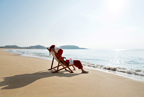 Santa Claus resting on deck chair at beach.