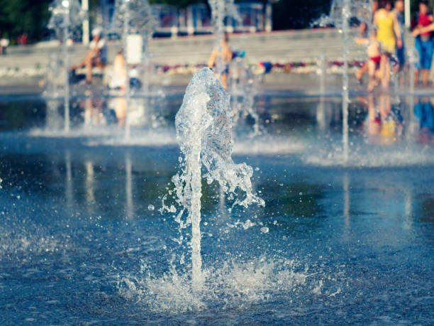 Children with parents cooling near dry fountain on main town square, close up view. Water streams splashing on granite coating. Summer cityscape. Selective soft focus. Blurred background niños con padres refrescando cerca de la fuente seca en la plaza principal de la ciudad, vista de cerca. corrientes de agua que salpican sobre el revestimiento de granito. paisaje urbano de verano. enfoque suave selectivo. fondo borroso.