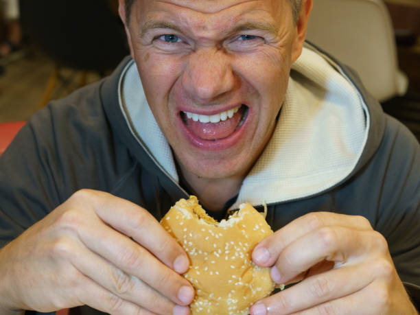 Man eating fast food in cafe and unsatisfied with it Portrait of handsome hungry man aged 40-50 y.o. eating fast food in city cafe. He bit off a piece of burger and looks at camera. He dissatisfaction with food. High resolution photography. brat burger stock pictures, royalty-free photos & images