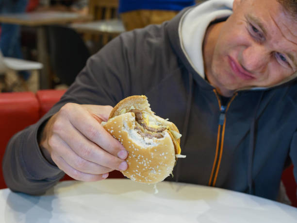 Man eating fast food in cafe and unsatisfied with it Portrait of handsome hungry man aged 40-50 y.o. eating fast food in city cafe. He bit off a piece of burger and looks at camera. He dissatisfaction with food. High resolution photography. brat burger stock pictures, royalty-free photos & images