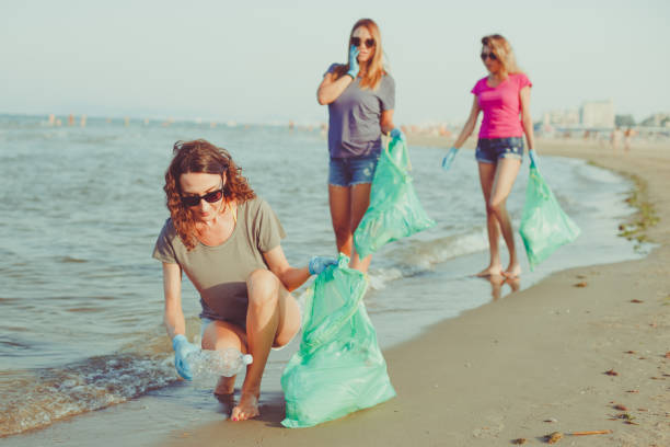 image of young women volunteers picking up (cleaning) plastic garbage on the beach - save the earth, ecology and plastic recycling concept - group of people women beach community imagens e fotografias de stock