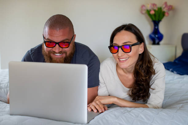 Happy couple relaxing at home with lap top, looking at the screen with orange blue light blocking glasses -  health, good sleeping and wellness concept for melatonin and circadian rhythm stock photo