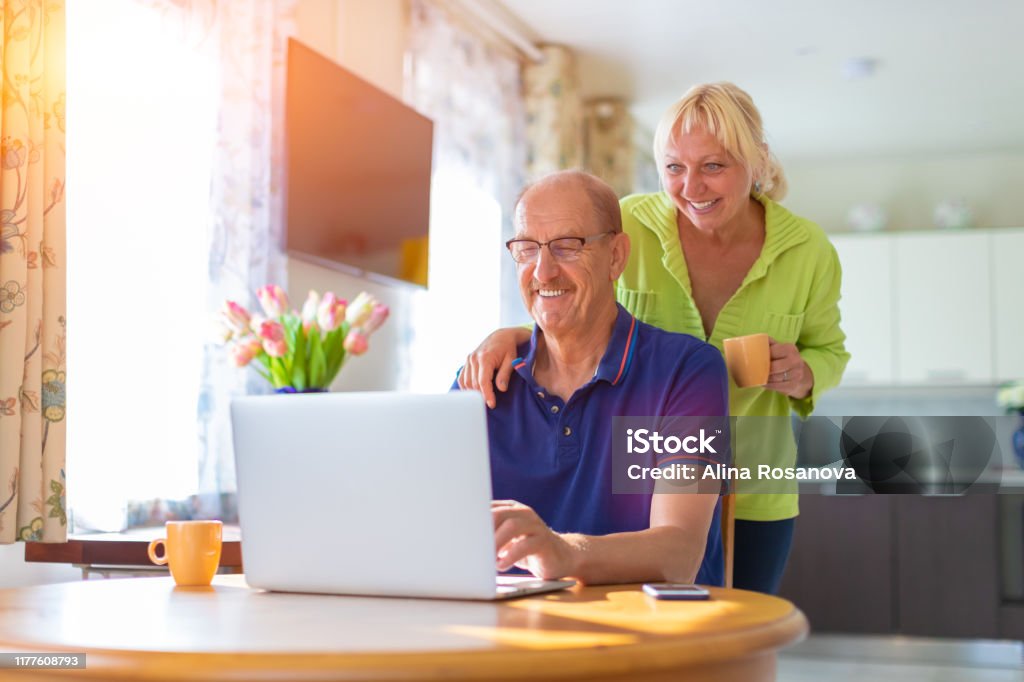 Senior couple looking at the lap top screen smiling and drinking coffee - elderly people video calling or talking by web camera - working at home, freelancing and having hobbies together concept Portrait of two people with computer on the table Netherlands Stock Photo