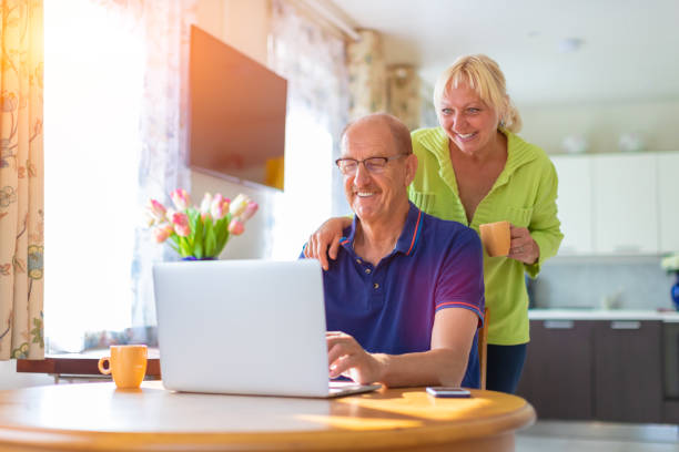 Senior couple looking at the lap top screen smiling and drinking coffee - elderly people video calling or talking by web camera - working at home, freelancing and having hobbies together concept stock photo