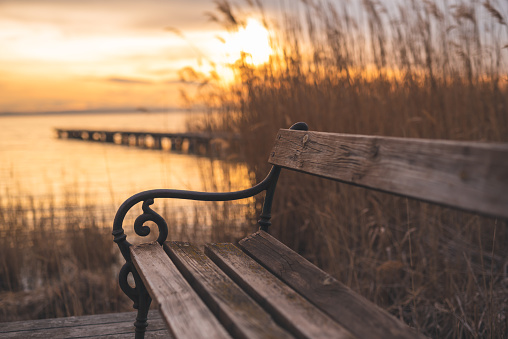 Autumnal sunset at the lake. Lakeshore with a bench and wooden walkway. In the background the footbridge, the reed and the setting sun. Autumn at the lake. Winter at the lake