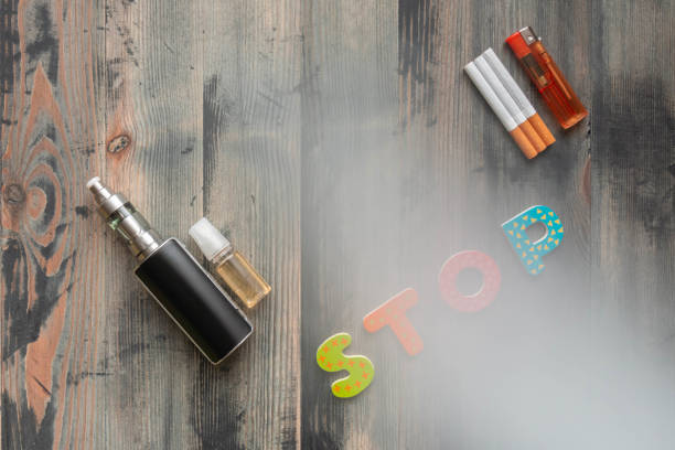 Flat lay of e-cigarette, e-liquid, tobacco cigarettes with lighter and letters stop on wooden background, covered smoke stock photo