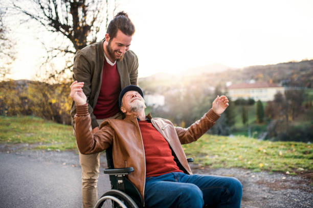 Young man and his senior father in wheelchair on a walk in town. A young man and his senior father in wheelchair on a walk in town at sunset. wheelchair photos stock pictures, royalty-free photos & images