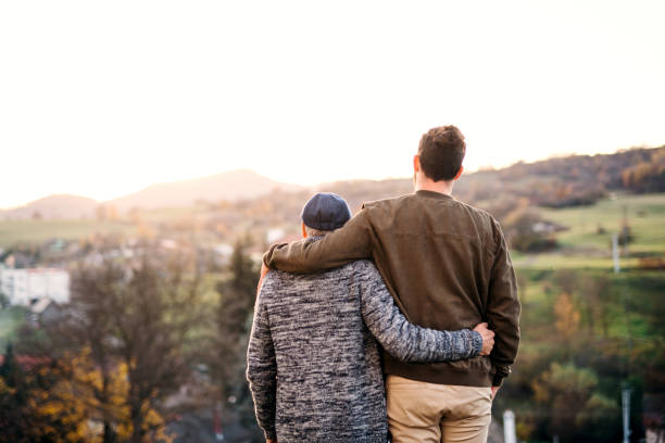 Rear view of senior father and his son on a walk in park, standing. Rear view of senior father and his son on a walk in park, standing arm in arm. locking-arms stock pictures, royalty-free photos & images