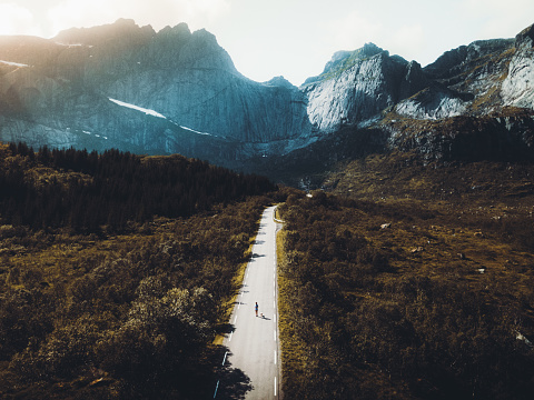 Drone photo of picturesque autumn sunset above Nusfjord on Lofoten Islands, Northern Norway - woman running with her dog on the abandoned road in from of big mountain and orange trees between road