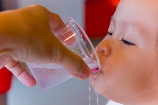 Adorable 7-month old baby boy sitting in highchair learning to drink water from a glass