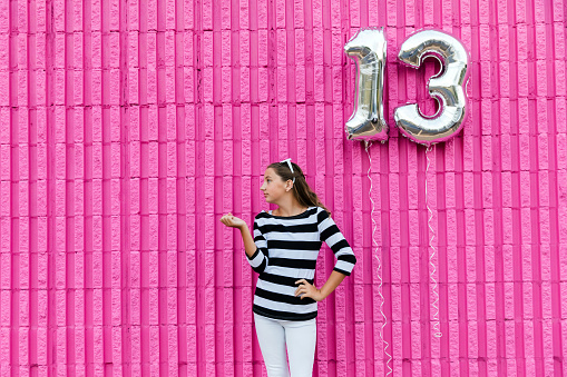 Cute caucasian girl with long hair in front of a hot pink wall outside wearing a striped shirt and sunglasses making teen girl poses with silver mylar helium balloons to celebrate becoming a teenager. Looking to the side with her hand up.