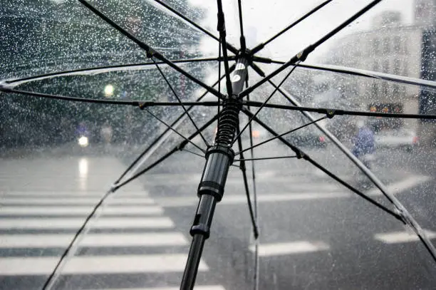 Open umbrella under light rain in the streets of a busy city. Clear parasol with black metal frame.