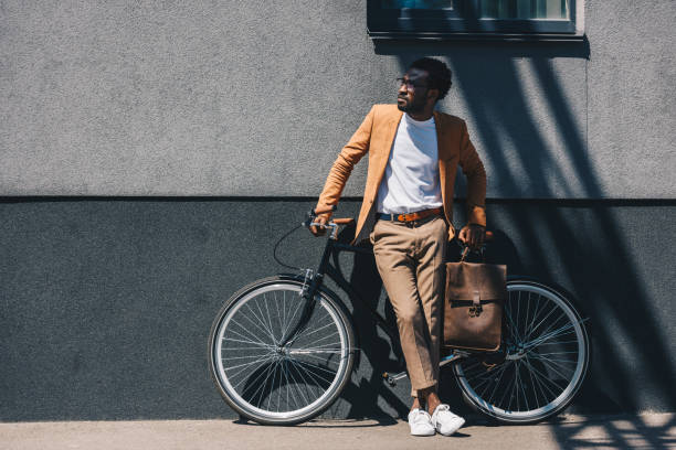 guapo hombre de negocios afroamericano de pie junto a la pared cerca de la bicicleta y mirando hacia otro lado - african descent cycling men bicycle fotografías e imágenes de stock