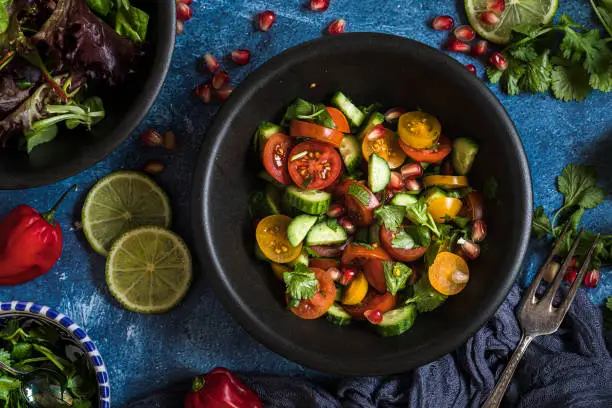 Fresh salad with tomato, cucumber and herbs, flat lay from above  The salad is in a black bowl and there are other bowls with ingredients around it. The table is decorated with lime, chili and cilantro.