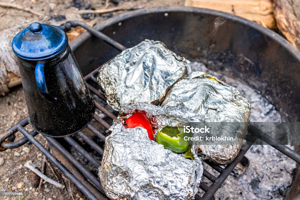 Blue tea water kettle and foil wrapped red green pepper vegetables on grill in fire pit at campground cooking dinner Cooking Stock Photo