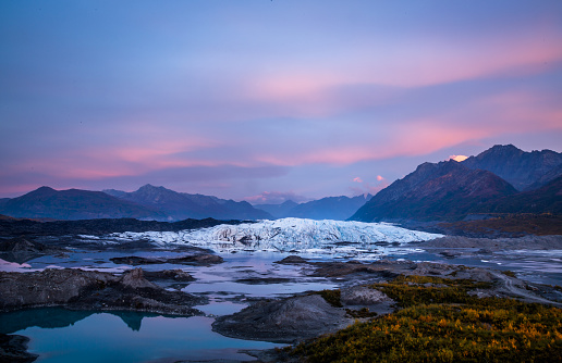Clouds pink and purple at sunset over the Matanuska Glacier in Alaska. Below the glacier a calm lake reflects the colors and leaves have started to turn yellow for Autumn.