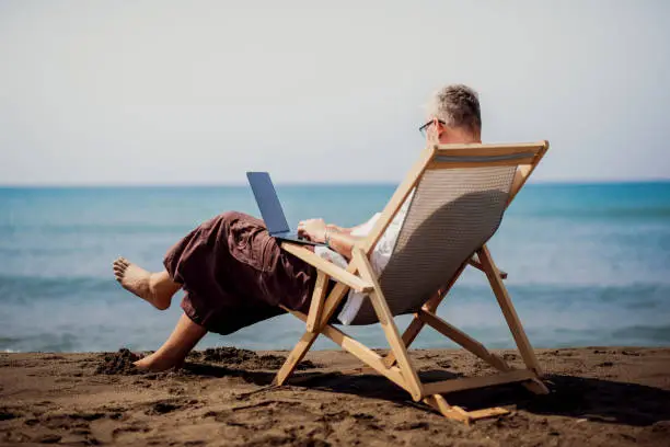 Gray-haired man is sitting next to the blue sea in a lounge chair and working on his laptop