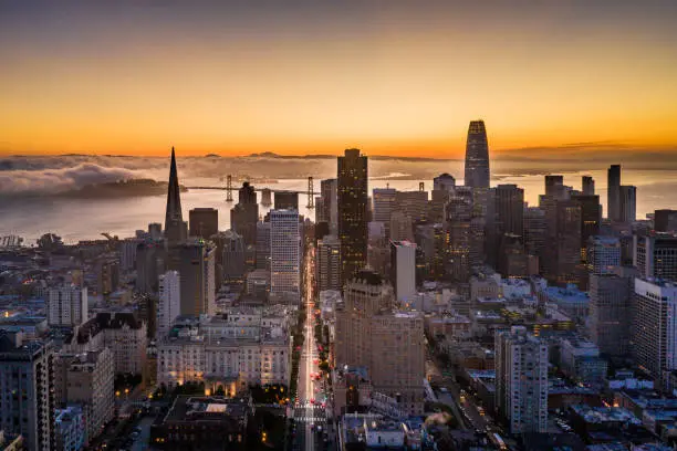 Aerial view of sunrise in San Francisco, California. View of Nob Hill, the financial district, iconic buildings and the well known California Street with the Bay Bridge at the end of the street. Well know buildings line the street. Treasure island and the San Francisco Bay have fog rolling across. Twinkling lights across the city skyline.