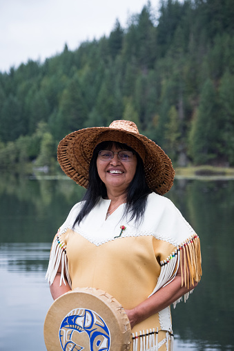 A First Nations woman Standing in the forest. She is dressed is full traditional clothing and is wearing a woven hat that she made. She is happy and smiling