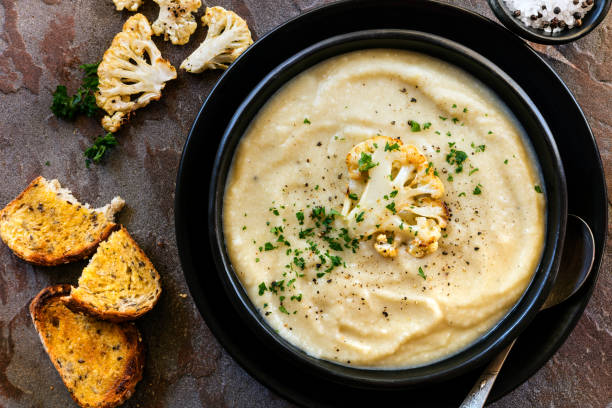 Cauliflower Soup in Rustic Black Bowl Top View Roasted cauliflower soup in rustic black bowl, with crusty sourdough toast.  Top view. soup stock pictures, royalty-free photos & images