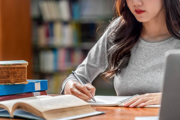 istock Closeup Asian young Student hand writing homework and using technology laptop in library of university or colleage with various book and stationary over the book shelf background, Back to school 1177789187