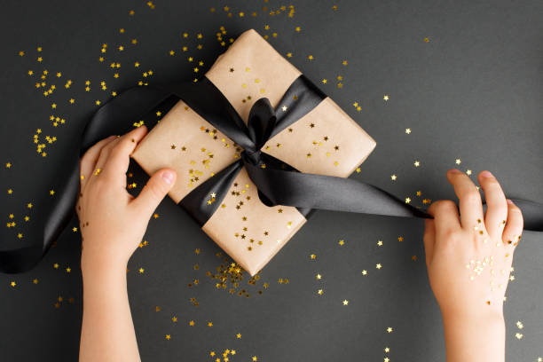 istock Gift box with black bow on black background with delicate gold star sparkles, kids hands packaging present magical holiday time, black friday sale concept 1177801039