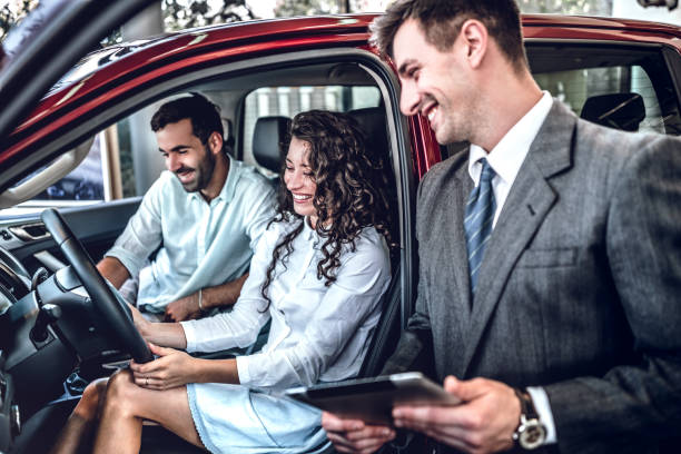 seller of prestigious car dealership telling characteristics of vehicle. young couple in love sitting in salon of liked car - professional dealer imagens e fotografias de stock