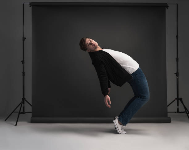 Young stylish man posing and dancing in the studio on a gray background. Dressed in a white T-shirt, blue jeans, black jacket and white sneakers. Model tests for men.