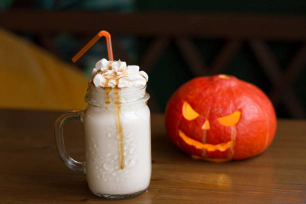 ホイップクリーム、マシュマロ、カボチャキャラメルシロップ、秋の秋のカボチャジャック-オランタンの隣の木製テーブルのオレンジストローとガラス瓶でミルクシェイク - glass autumn halloween food ストックフォトと画像