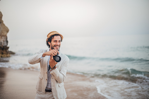 Handsome, bearded man with hat and white suit taking photos on the beach.