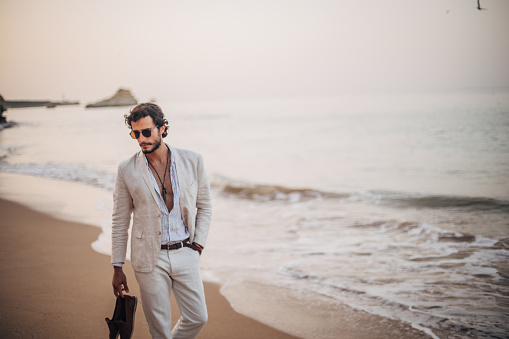 Handsome, modern man in casual, white suit, holding his shoes and taking a walk on the beach.