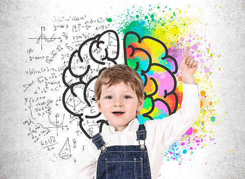 Adorable little boy in jeans overalls pointing up standing near concrete wall with colorful brain sketch and science formulas. Concept of creative thinking and education