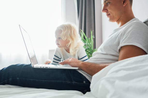 istock Young dad sits on bed with children using the computer together at home. Father working on laptop while his daughter looking on screen 1177837702