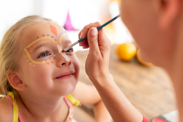 Young mother painting daughters face for Halloween party. Halloween or carnival family lifestyle background. Face painting and dressing up. stock photo