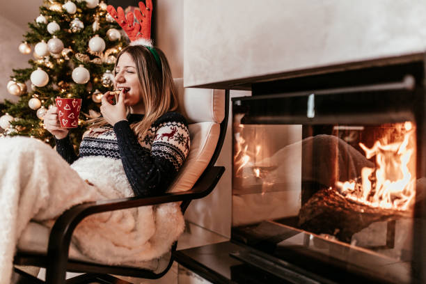 Young beautiful woman celebrating New Year or Cristmass at home alone. She sitting next to the modern fireplace, eating cookies, drinking tea and enjoying in peace.