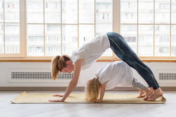 Photo of Young mother doing yoga with 3-years girl in front of window. Downward facing dog asana