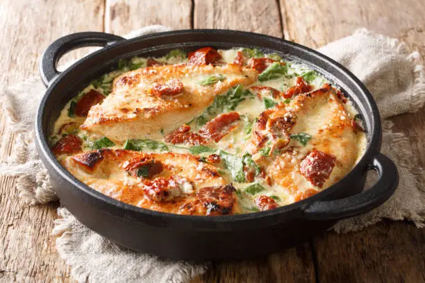 Photo of Main course baked chicken with sun-dried tomatoes and spinach in cheese sauce close-up in a pan. horizontal