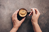 Female hands holding spoon and stirring hot coffee on a dark brown rustic background. Top view, flat lay.