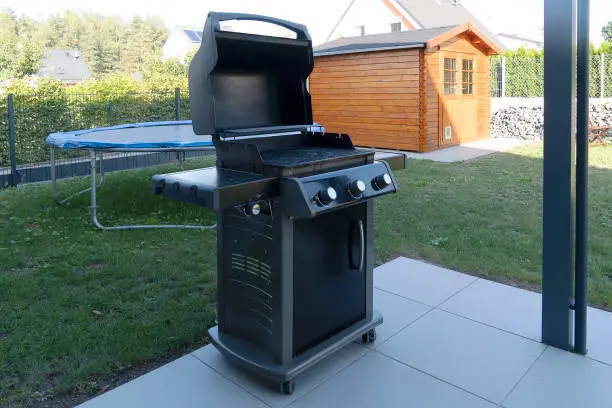 A large black gas grill stands on the street on the lawn near the house. Street cooking.