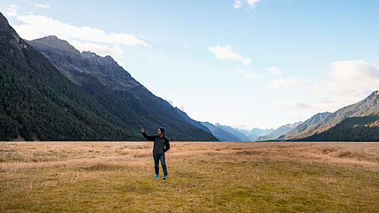 A tourist selfie with the smartphone to capture the beautiful landscape scenic view of Ellington Valley, the viewpoint along the road to Milford sound, the place have vast meadow with mountain ranges.