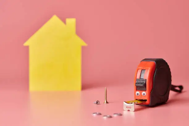 Photo of Metal tape measure funny concept. House renovation. Home repair and redecorated concept. Yellow house shaped figure on pink background.