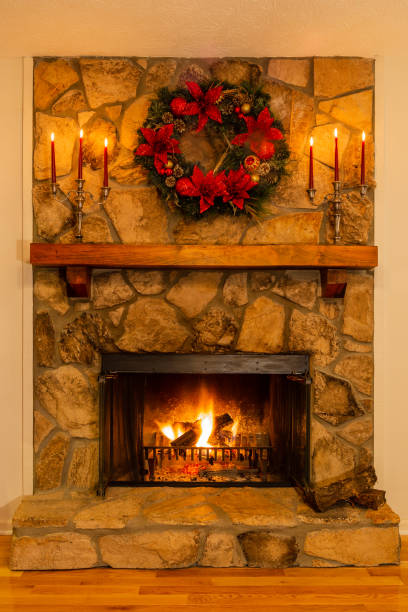 Stone fireplace decorated with a green and red wreath and red candles burning. Holiday decorated fireplace with a blazing fire, candles and a Christmas wreath. poinsettia christmas candle flower stock pictures, royalty-free photos & images