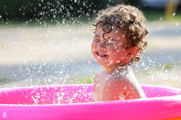 Baby boy in plastic swimming pool playing with water and splashing drops outside.