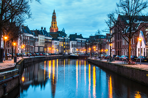 The photo is shot along a canal at night in Groningen with the Aa-Kerk church and historic center visible in the background. The street lights are reflected in the river. The sky is blue with copy space.