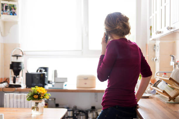 una giovane donna a casa in cucina sta usando un telefono, mentre guarda fuori dalla finestra - rear view women back back of head foto e immagini stock