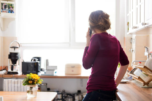 A young woman at home in the kitchen is using a telephone, while looking out of the window A woman is at home in her apartment making a phone call. She’s alone. In the background visible is a bright lit window with natural light and copy space. back of head photos stock pictures, royalty-free photos & images