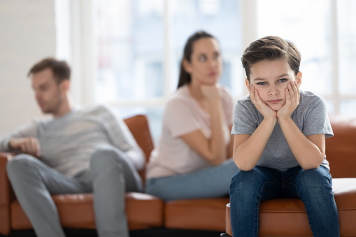 Upset little preschool child boy looking at camera feel sad hurt about parents fights conflicts sit on sofa at home, depressed school kid son suffer from family argument, children and divorce concept