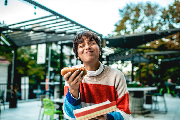 Young woman eating hot dog on the street a young girl holds a fast food meal in her hand while she laughs and looks at the camera eating stock pictures, royalty-free photos & images