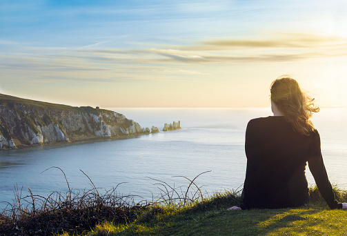 A female tourist is sitting on the edge of a cliff overlooking the famous chalk stone Needles and 19th century lighthouse on the coastline of the Isle of Wight, a small island off the south coast of England, UK. The nature background photo was taken around sunset.