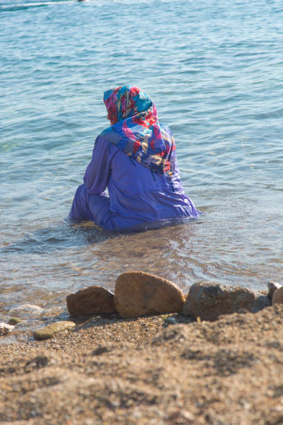 穆斯林婦女正坐在海邊。穆斯林婦女穿著藍色布基尼（伊斯蘭婦女泳衣）在海上背景。布林基尼的其他名字是布卡或雜湊馬。 - 回教泳裝 圖片 個照片及圖片檔